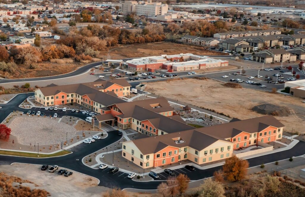 Aerial view of a suburban housing development with tan and brick townhomes, curved streets, and an empty lot amid autumn-colored trees.